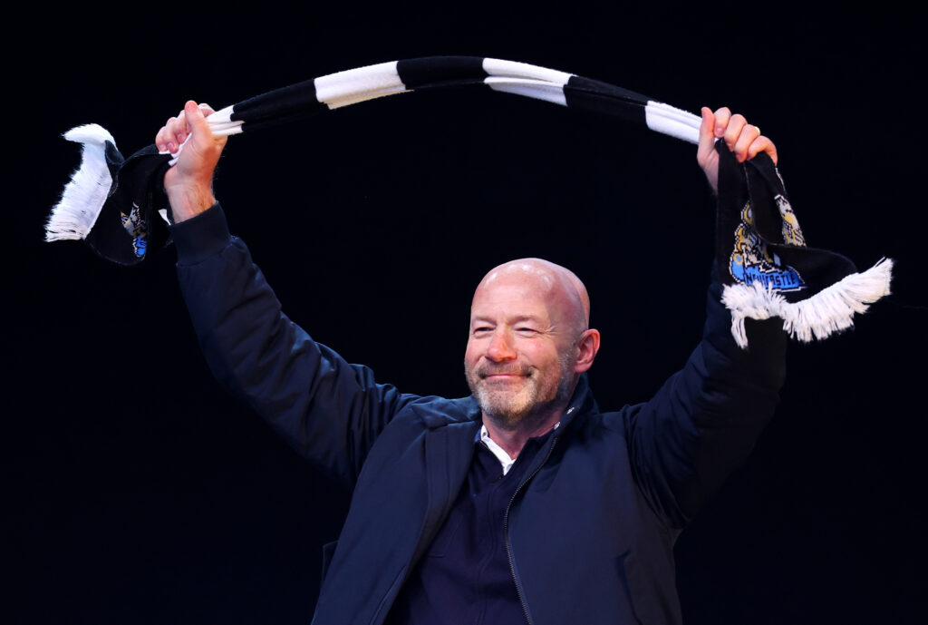 Alan Shearer holds a Newcastle United scarf in the air.